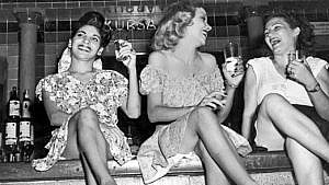 一张黑白照片，上面是三个穿着裙子的年轻女子，坐在酒吧里，拿着鸡尾酒，玩得很开心